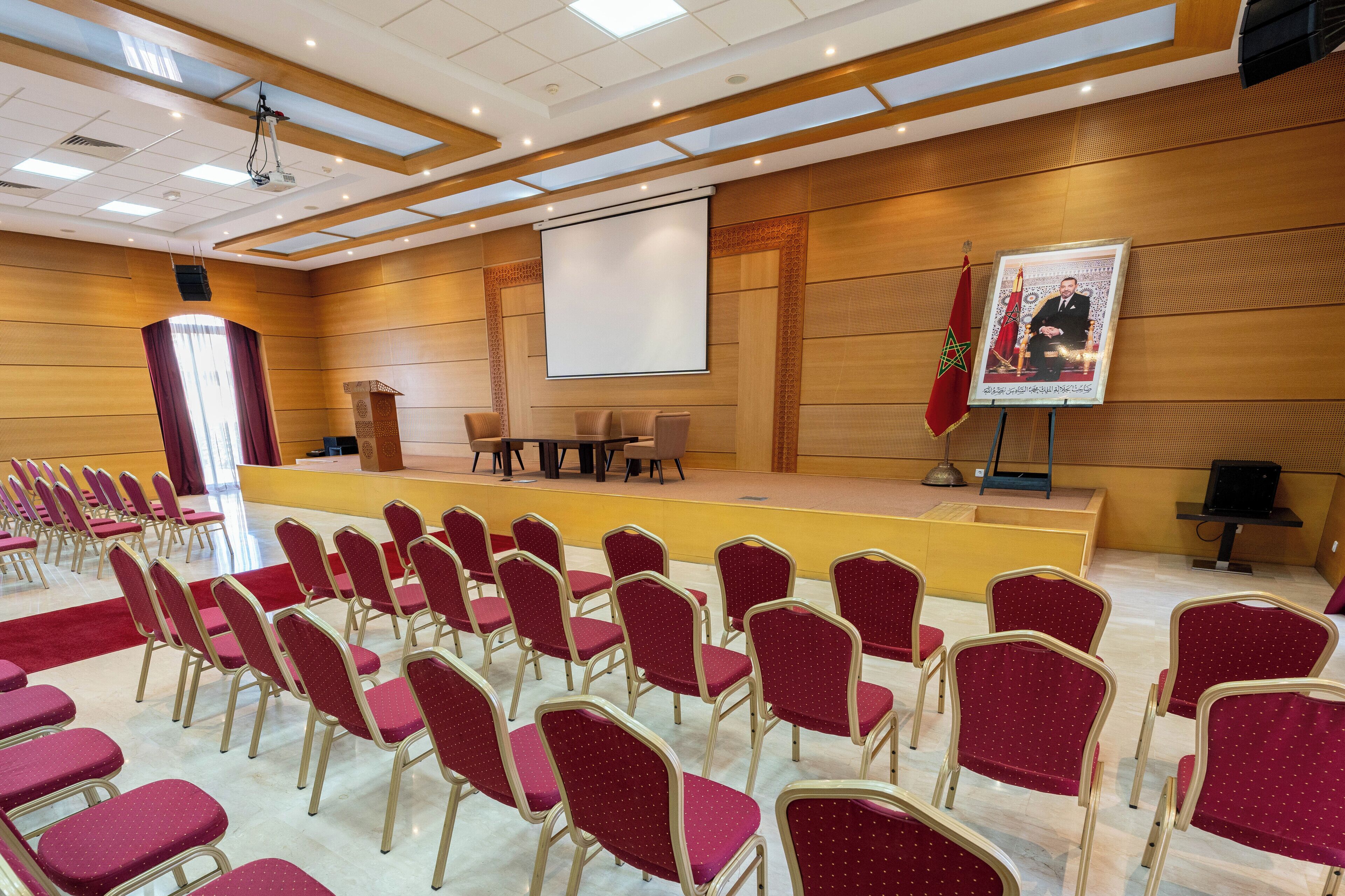 A conference room with rows of red chairs, a stage with a podium, and a large screen.