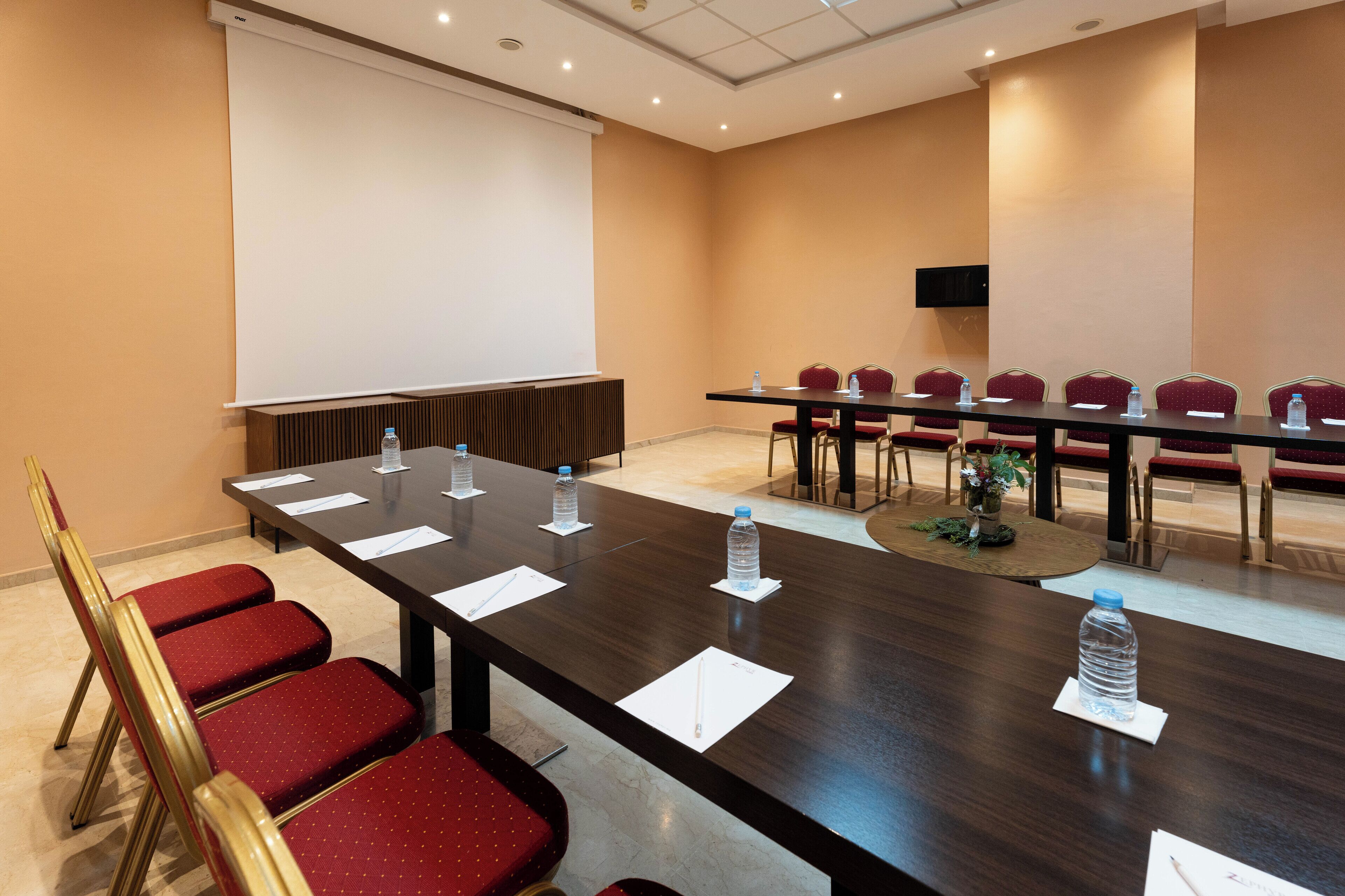 A conference room with a long table, red chairs, water bottles, and a projector screen.