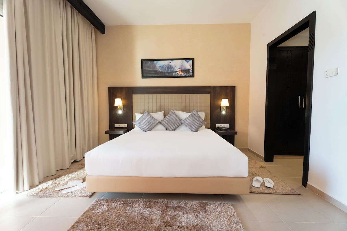 A large bed with a headboard, two bedside tables with lamps, a framed picture on the wall, and a pair of slippers on the floor.