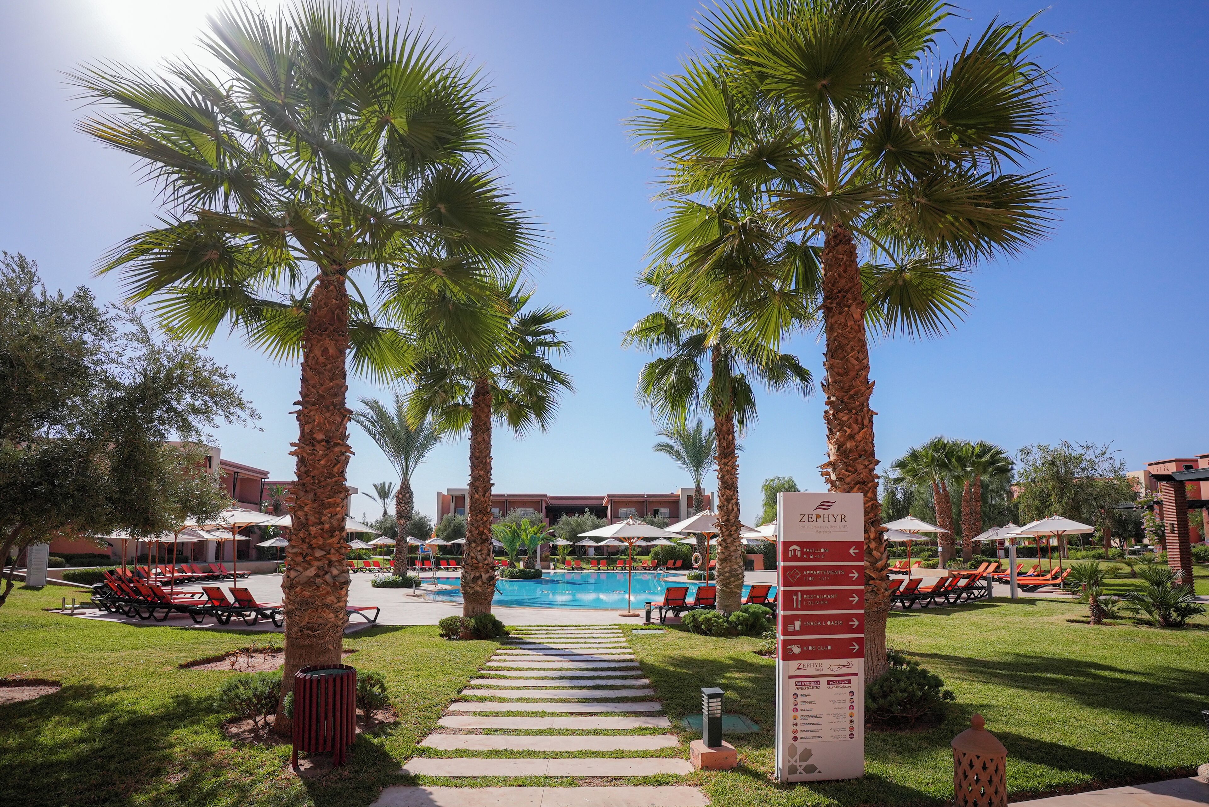 A resort pool area with palm trees, lounge chairs, and a sign indicating amenities.