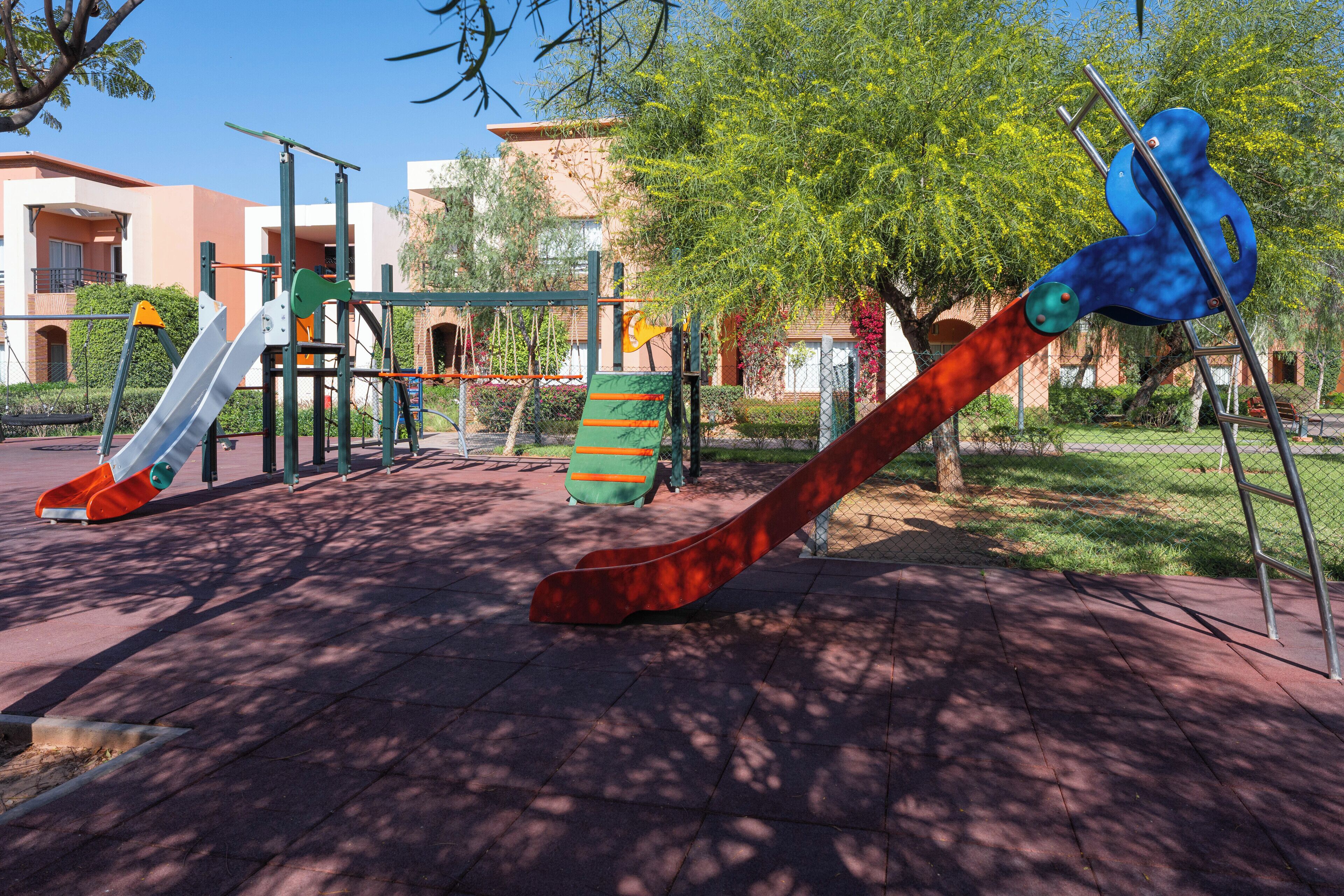Ein Spielplatz mit verschiedenen Spielgeräten, darunter Rutschen und Schaukeln, vor einer Kulisse aus Gebäuden und Bäumen.