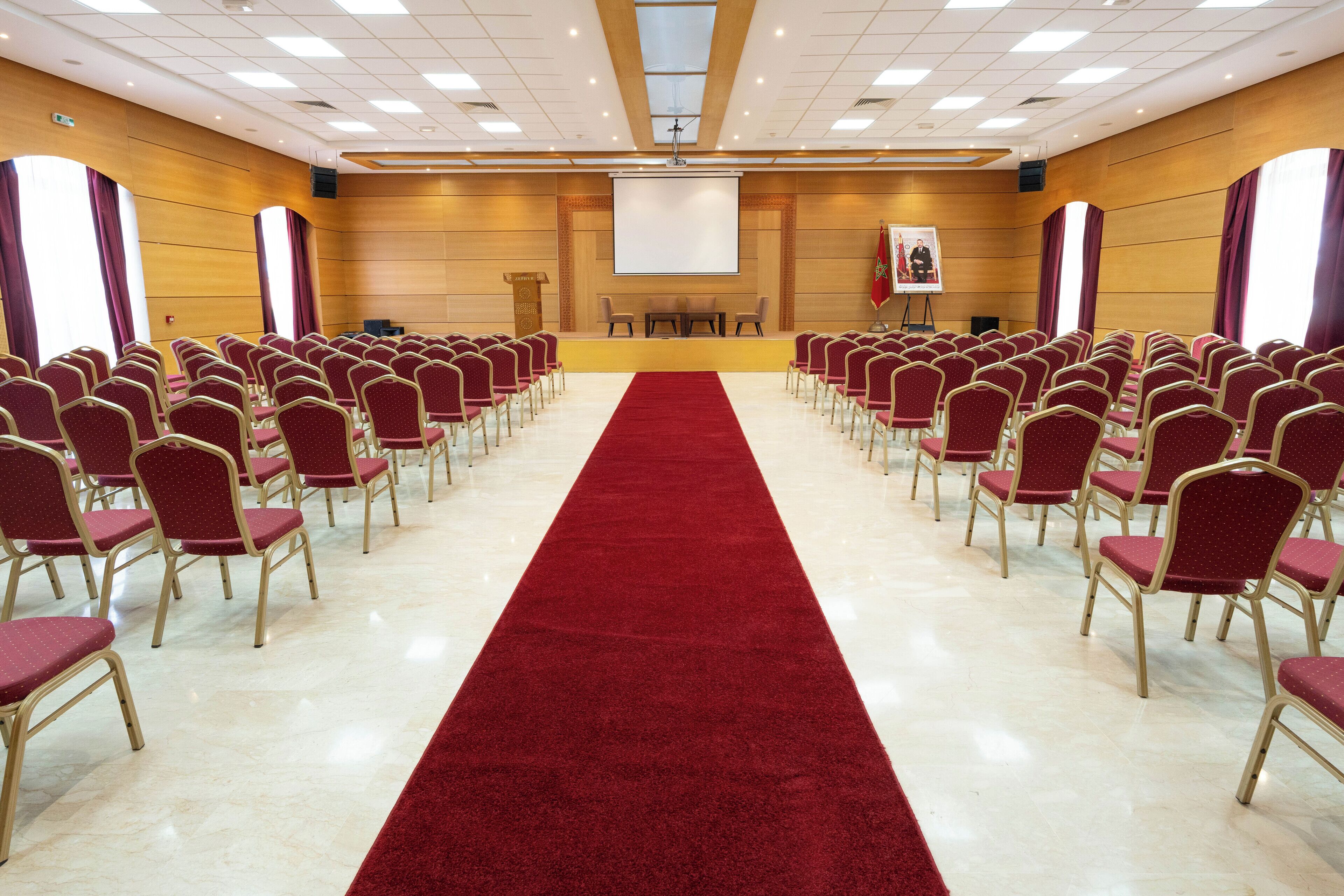 A conference room with rows of red chairs, a red carpet, and a projector screen at the front.