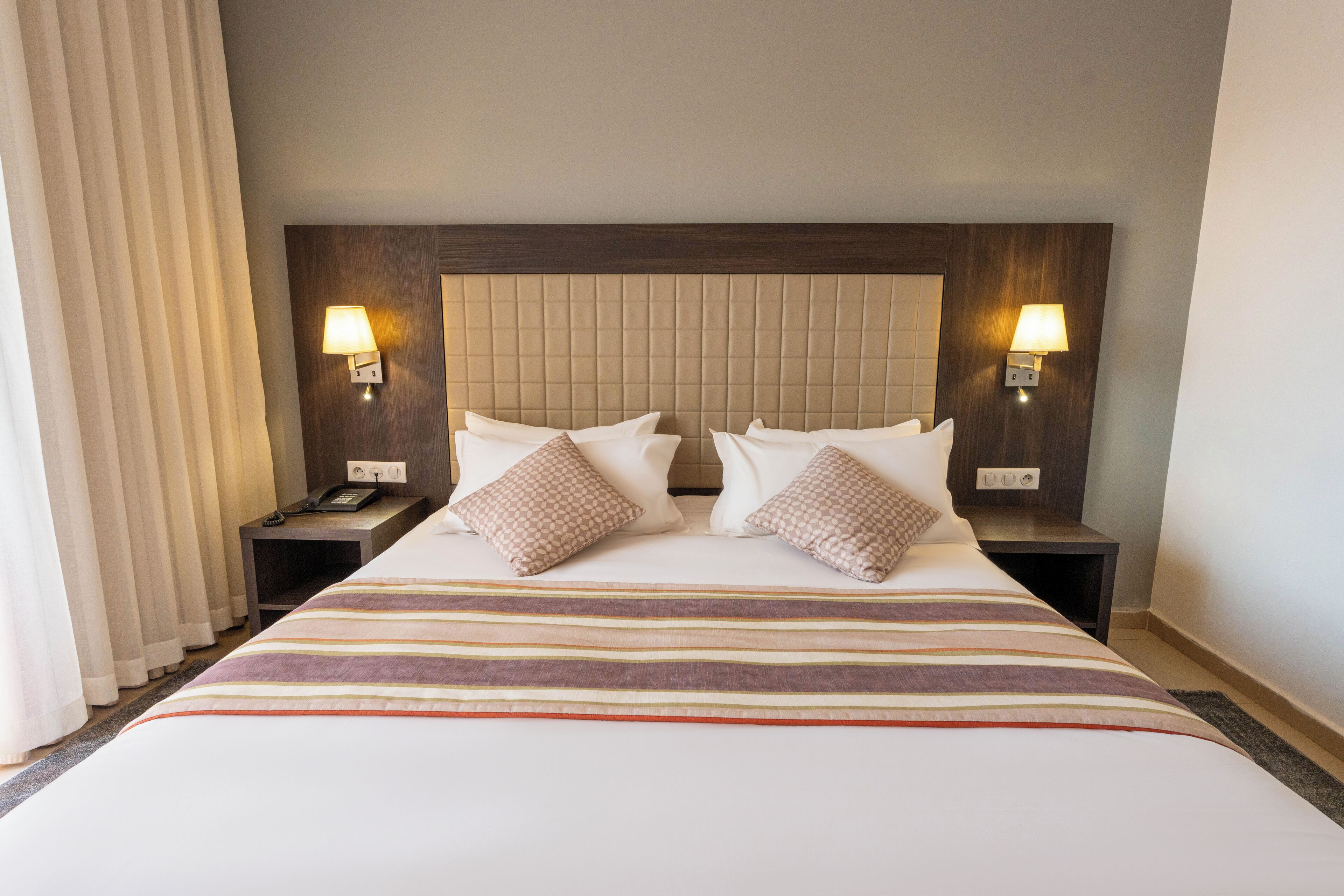 Une chambre d’hôtel avec un grand lit, deux tables de chevet, une tête de lit et deux lampes fixées au mur.