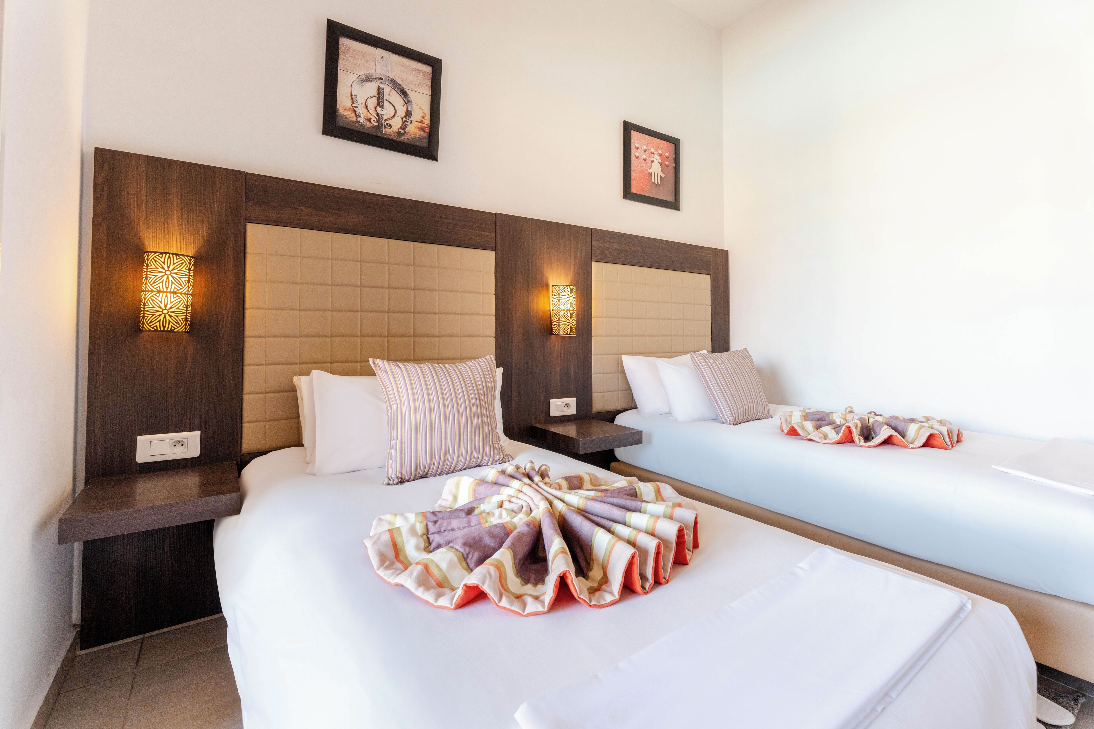 A hotel room with two beds, a wooden headboard, wall-mounted lamps, and a framed picture on the wall.