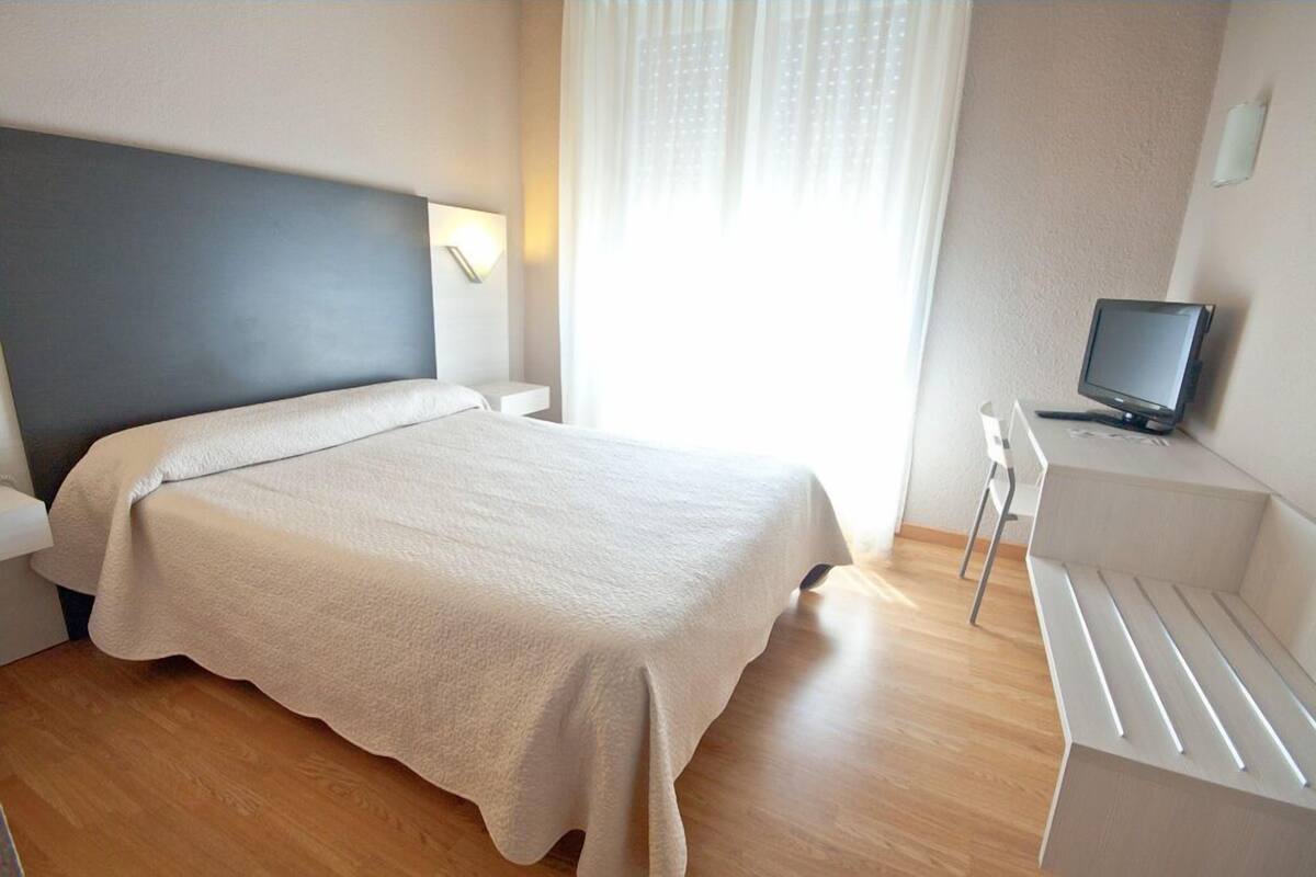 A hotel room with a bed, a desk with a computer, and a window with curtains.