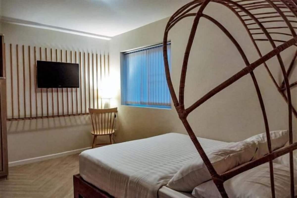 A bedroom with a bed, a TV mounted on the wall, a window with blinds, and a chair.