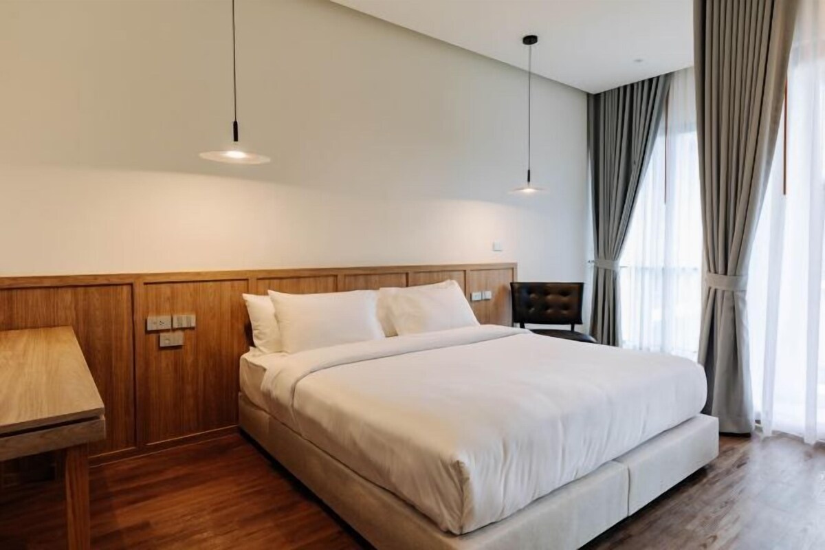 A hotel room with a large bed, wooden headboard, bedside tables, and a view of the outdoors through the window.
