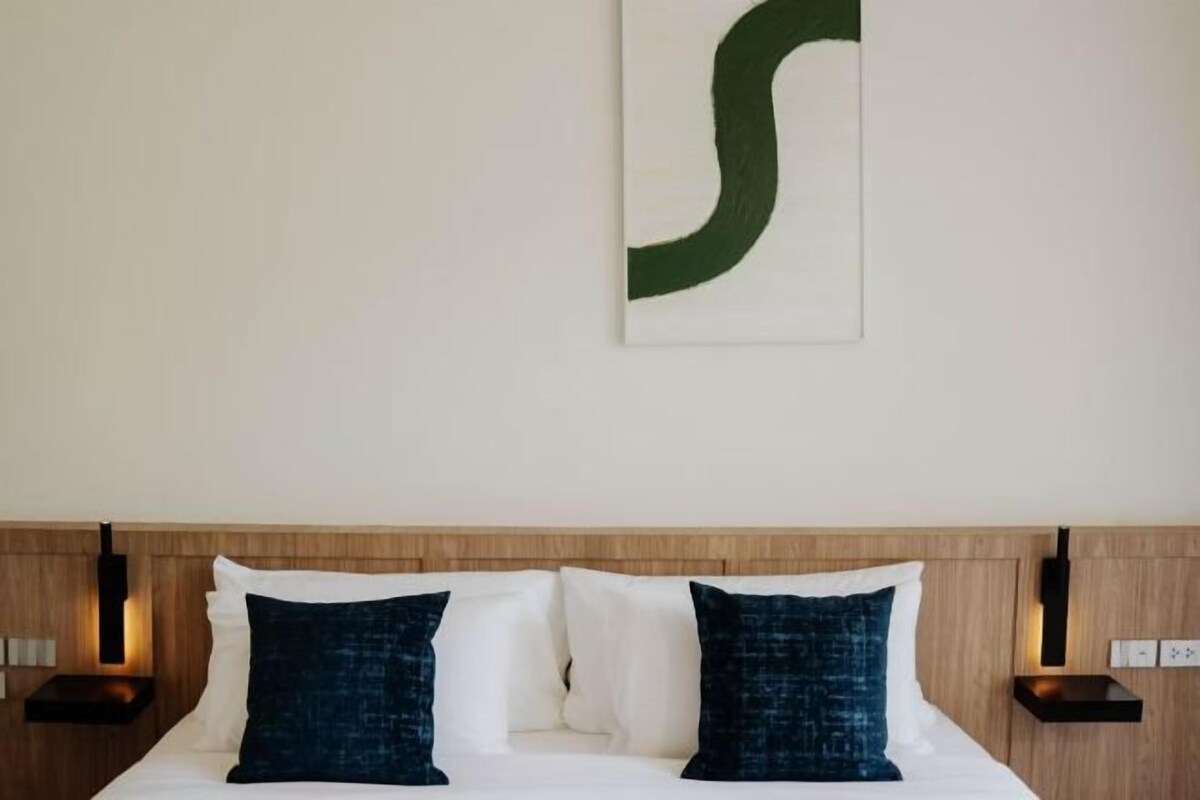A hotel room with a wooden headboard, two white pillows, two dark blue pillows, and a wall-mounted artwork.