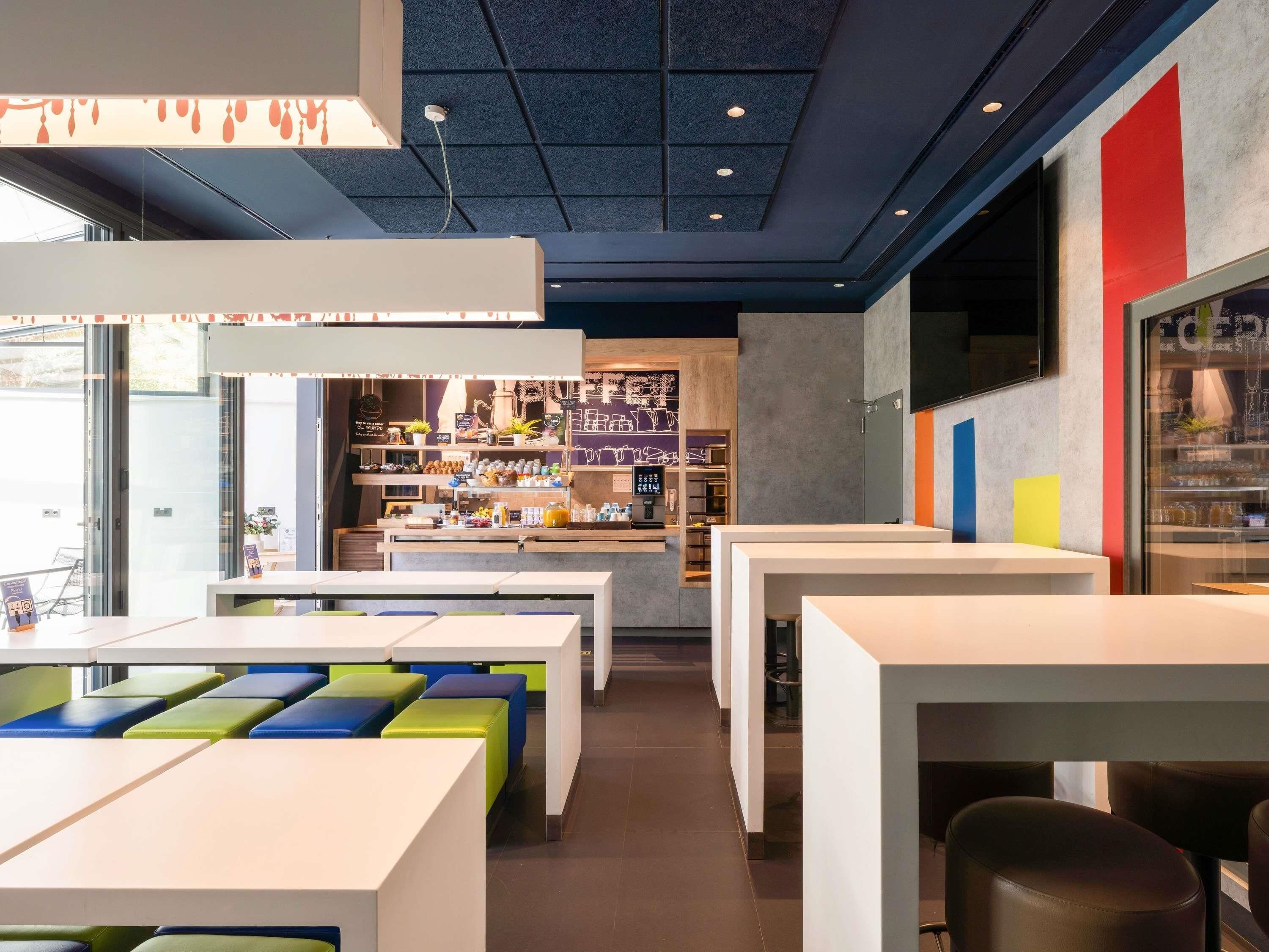 L’intérieur d’un café moderne, avec des tables blanches, des sièges aux couleurs vives et un comptoir bien visible où sont exposés divers articles.