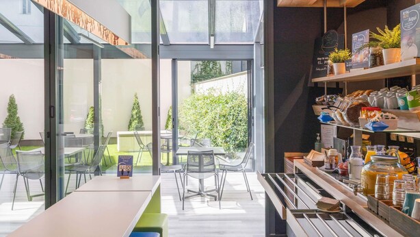 Un café moderne avec un mur de verre, des places assises en plein air et un bar où l’on sert le petit-déjeuner, avec un choix varié de plats.