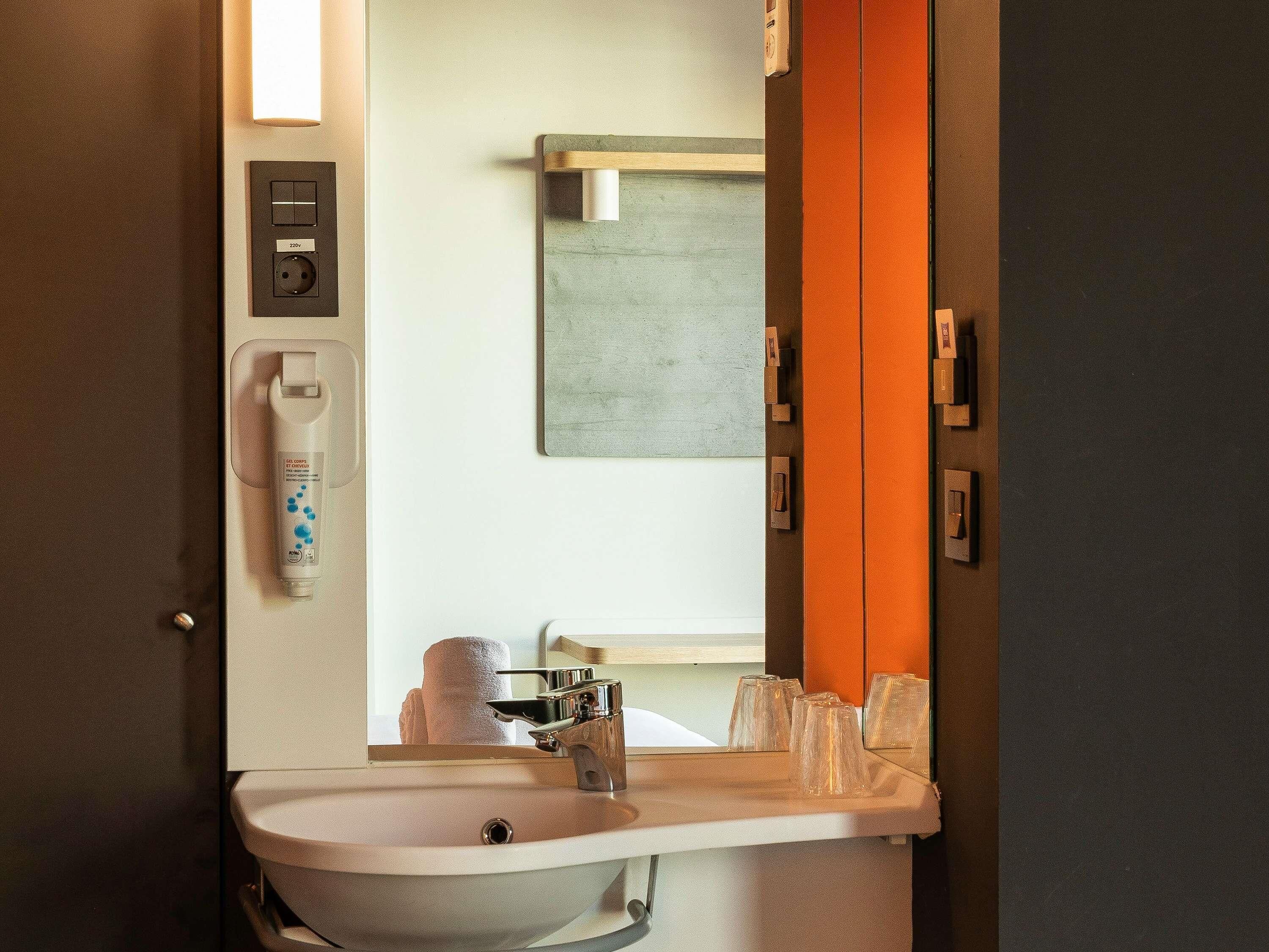 A bathroom with a sink, mirror, towel dispenser, and a toilet visible in the background.