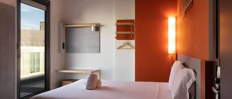 A hotel room with a bed, a desk, a hanging light, and a view through a window.