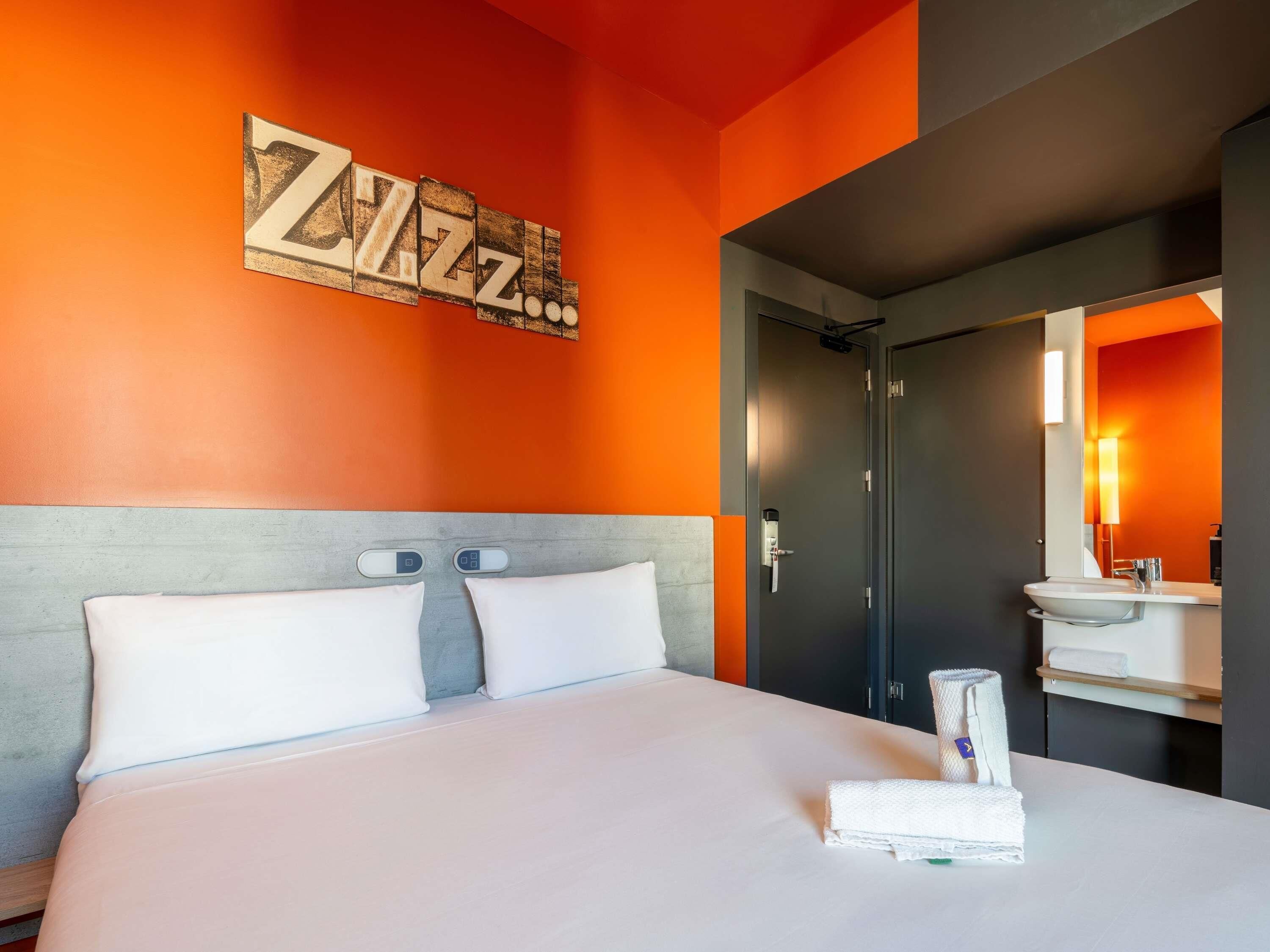 Habitación de hotel con cama, una obra de arte colgada en la pared, baño con lavamanos y espejo, y una puerta.