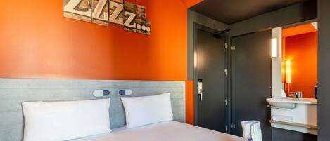 Habitación de hotel con cama, una obra de arte colgada en la pared, baño con lavamanos y espejo, y una puerta.