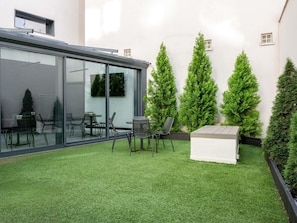 Une terrasse extérieure moderne, dotée d’un gazon synthétique, d’une véranda en verre et de chaises disposées avec soin.
