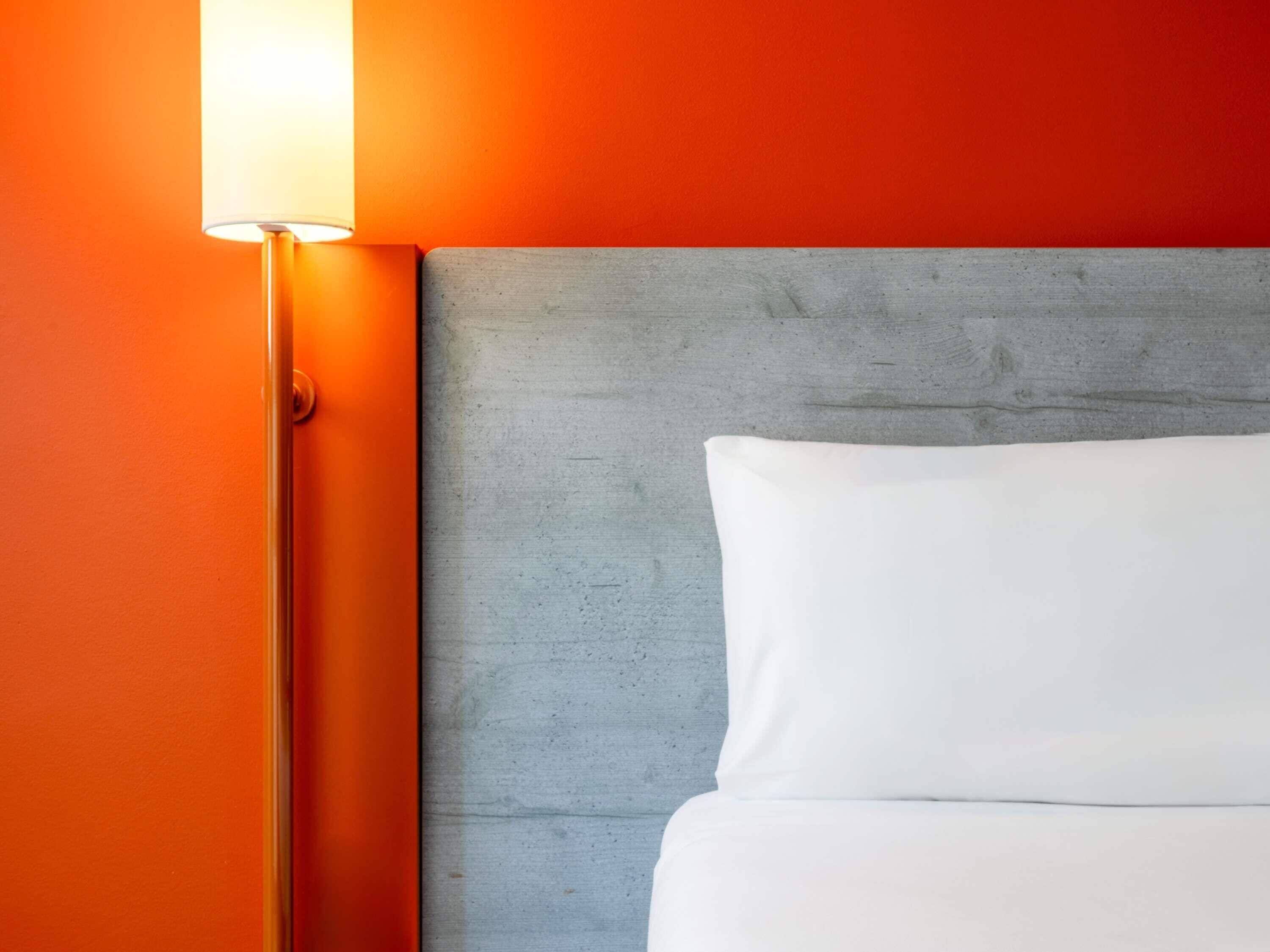 A bed with a grey headboard, a white pillow, and an orange wall.