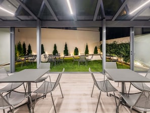 Un espacio interior moderno con mesas y sillas, un muro de cristal hacia un jardín y un techo con tragaluces.