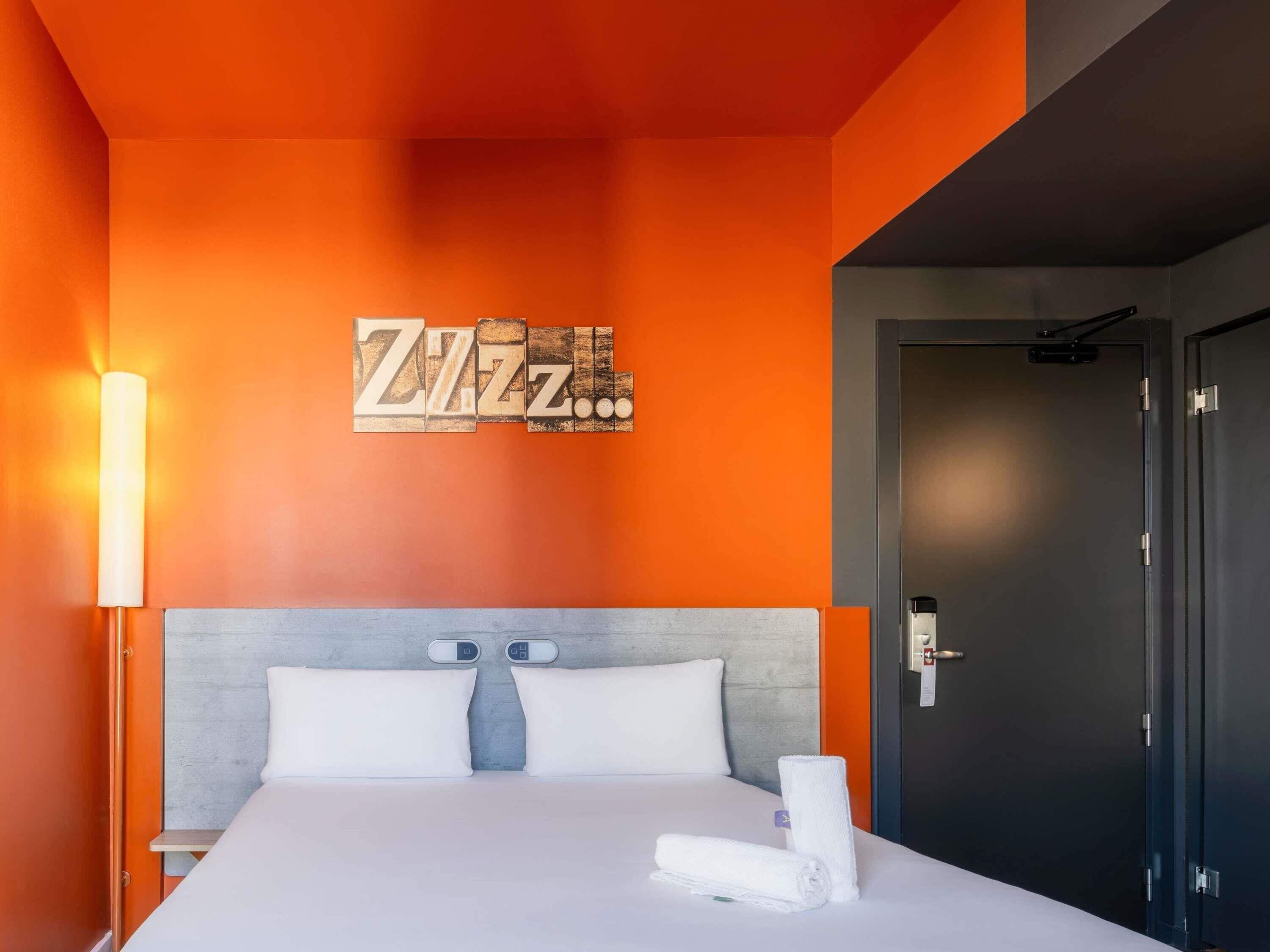 A hotel room with a bed, a wall clock, a door, and a wall art piece.