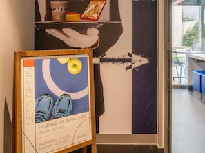 Une affiche encadrée représentant une personne tenant un plateau, un panneau indiquant les heures du petit-déjeuner, et une vue d’un intérieur moderne avec une table et des chaises.