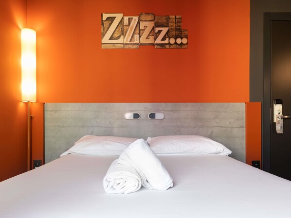 Habitación de hotel con cama, luz empotrada en la pared, termostato y puerta con cerradura.