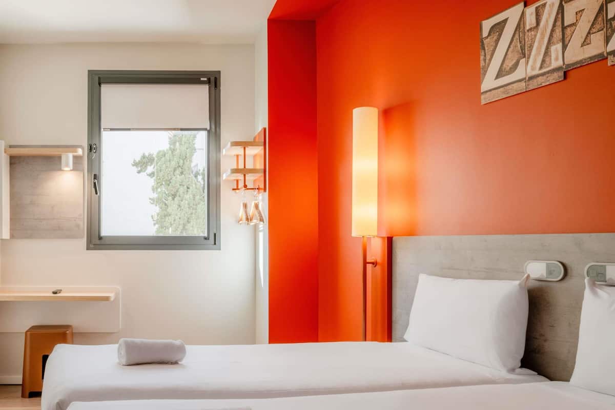 Une chambre d’hôtel avec deux lits, une fenêtre, un bureau et un mur d’accent de couleur rouge.