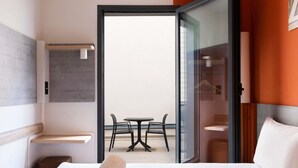 Une chambre d’hôtel comprenant un lit, une petite table, deux chaises, une porte vitrée donnant sur un balcon extérieur avec une table et des chaises, et un porte-serviettes fixé au mur.