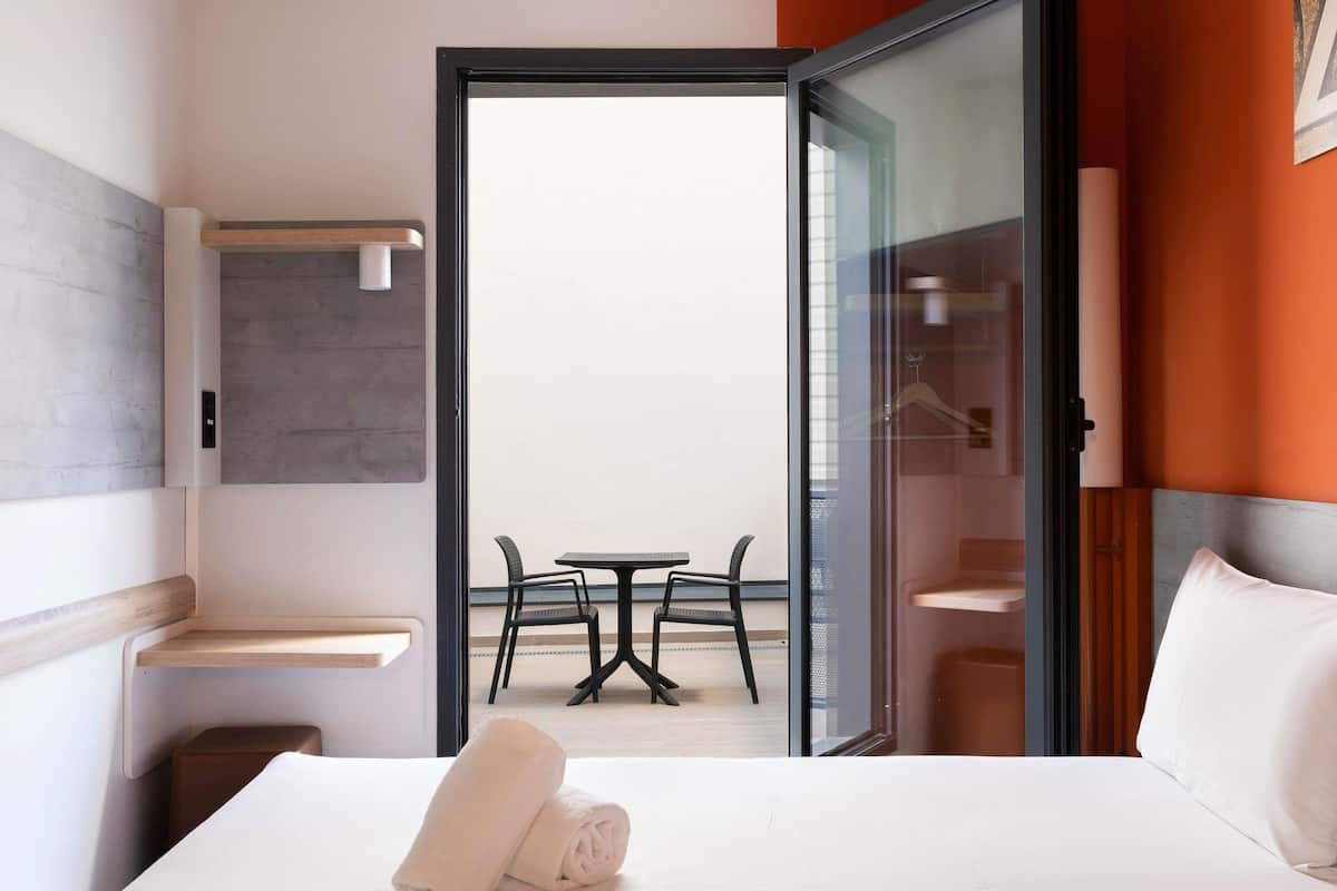 Une chambre d’hôtel comprenant un lit, une petite table, deux chaises, une porte vitrée donnant sur un balcon extérieur avec une table et des chaises, et un porte-serviettes fixé au mur.