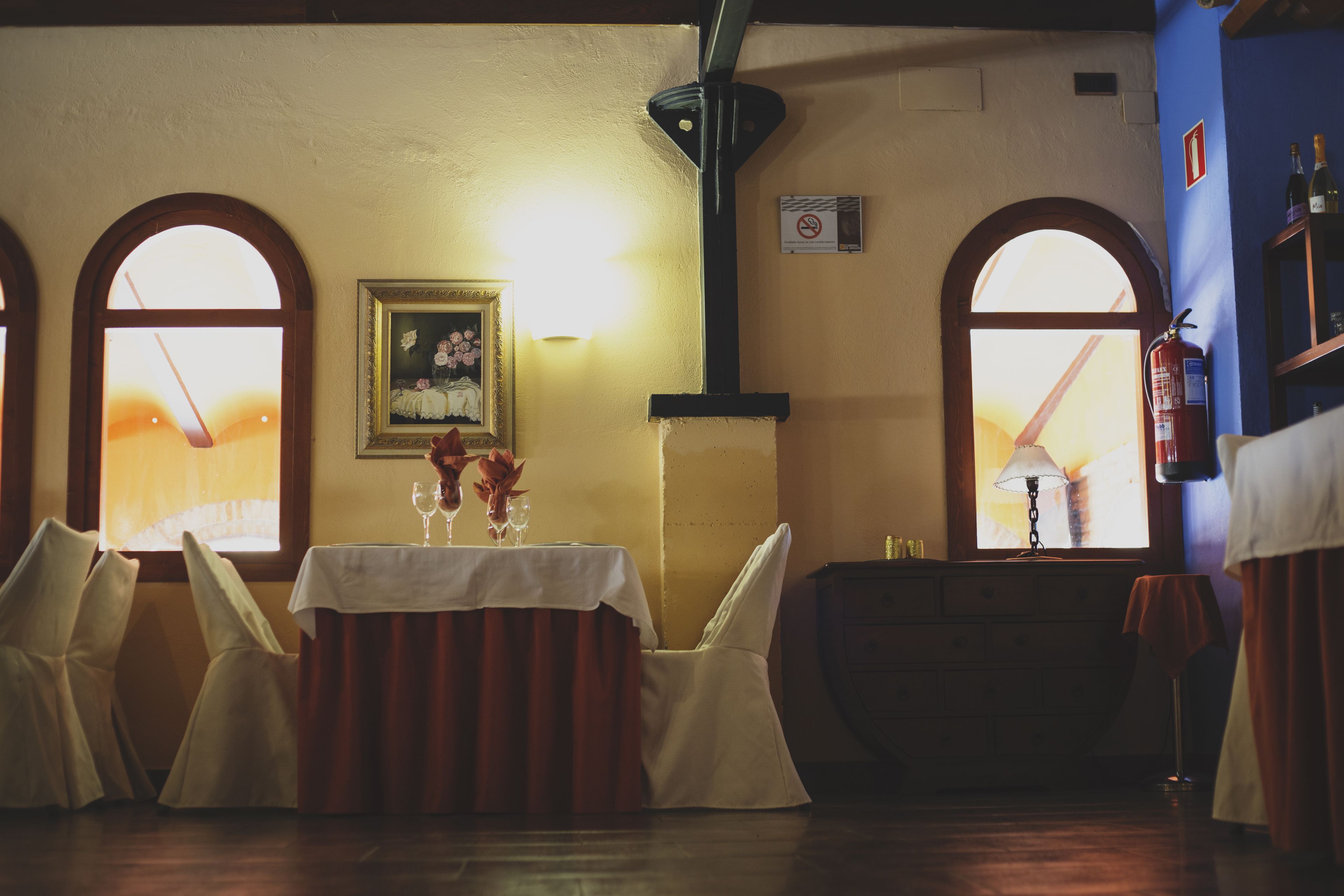 A dining area with a table set for four, two windows with arched tops, a framed picture on the wall, and a fire extinguisher mounted on the wall.