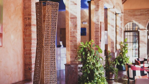 A brick interior with a tall, woven column, potted plants, and checkered chairs. - Hotel Restaurante La Bodega (La Almunia de Dona Godina)