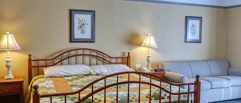 Una cama bien hecha con colcha, dos lámparas de noche, una mesita de noche, un sofá gris y dos cuadros con motivos florales en la pared.