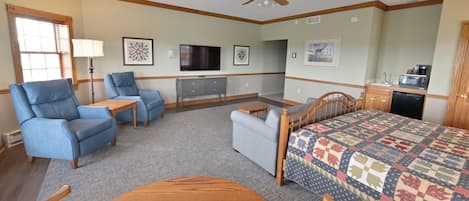 A hotel room with a bed, two armchairs, a desk, a television, a small table, and a kitchenette.