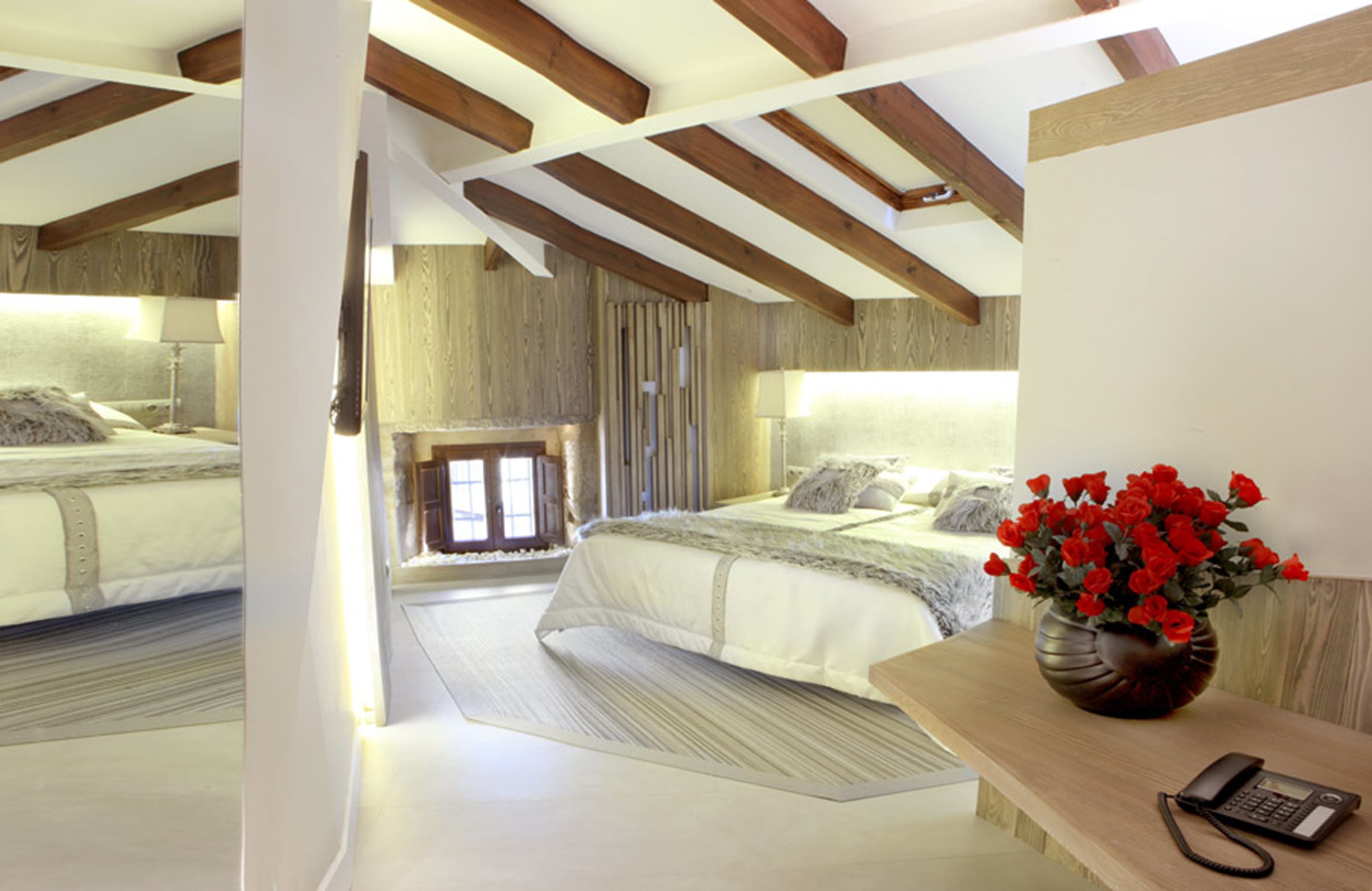 Un dormitorio con una cama grande, techo de madera y un florero con flores rojas sobre una mesita de noche.