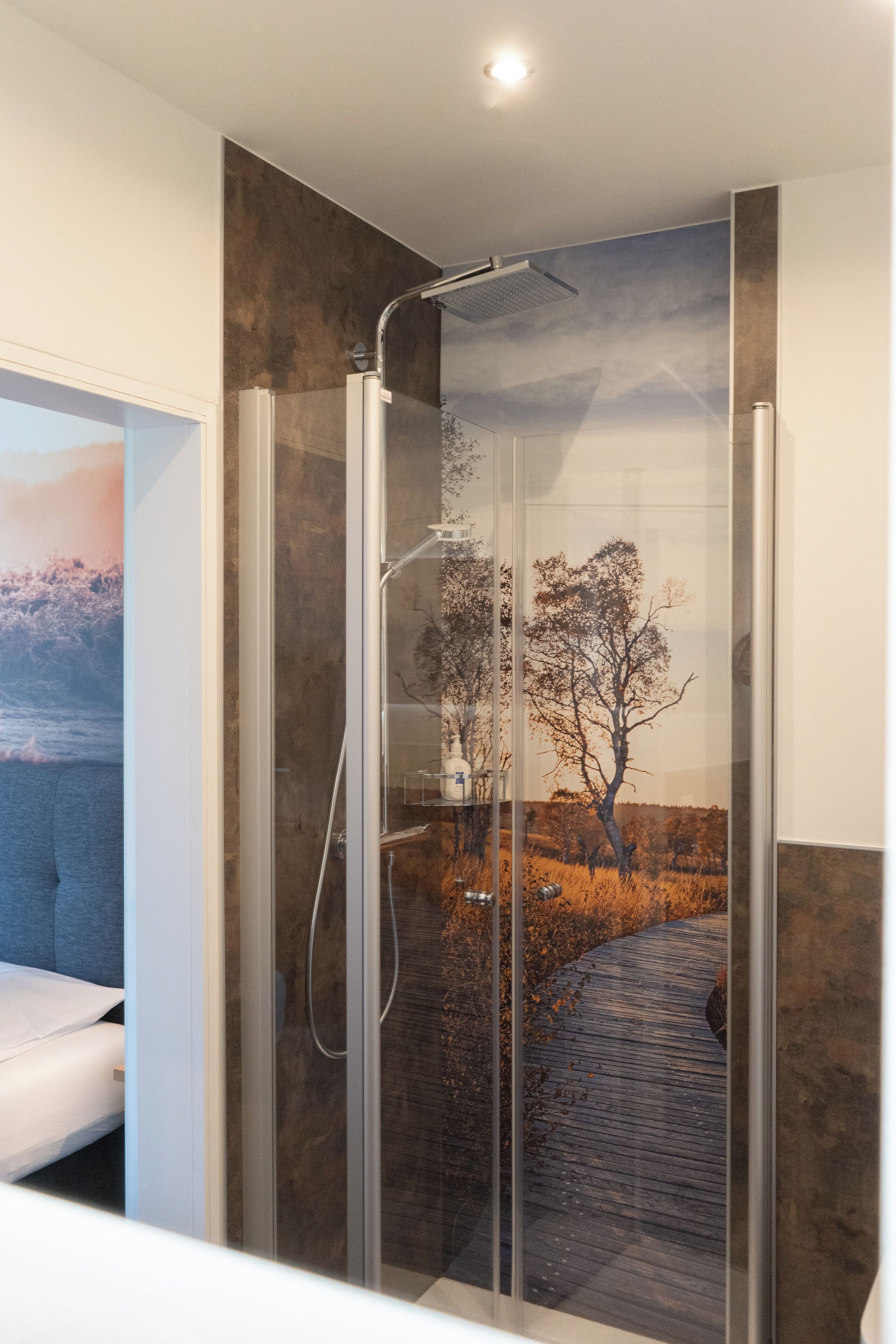 A modern bathroom with a glass-enclosed shower, a bed, and a scenic wall mural.