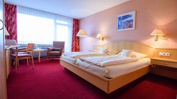 A hotel room with a large bed, two bedside tables, a desk with a chair, and a window with curtains.