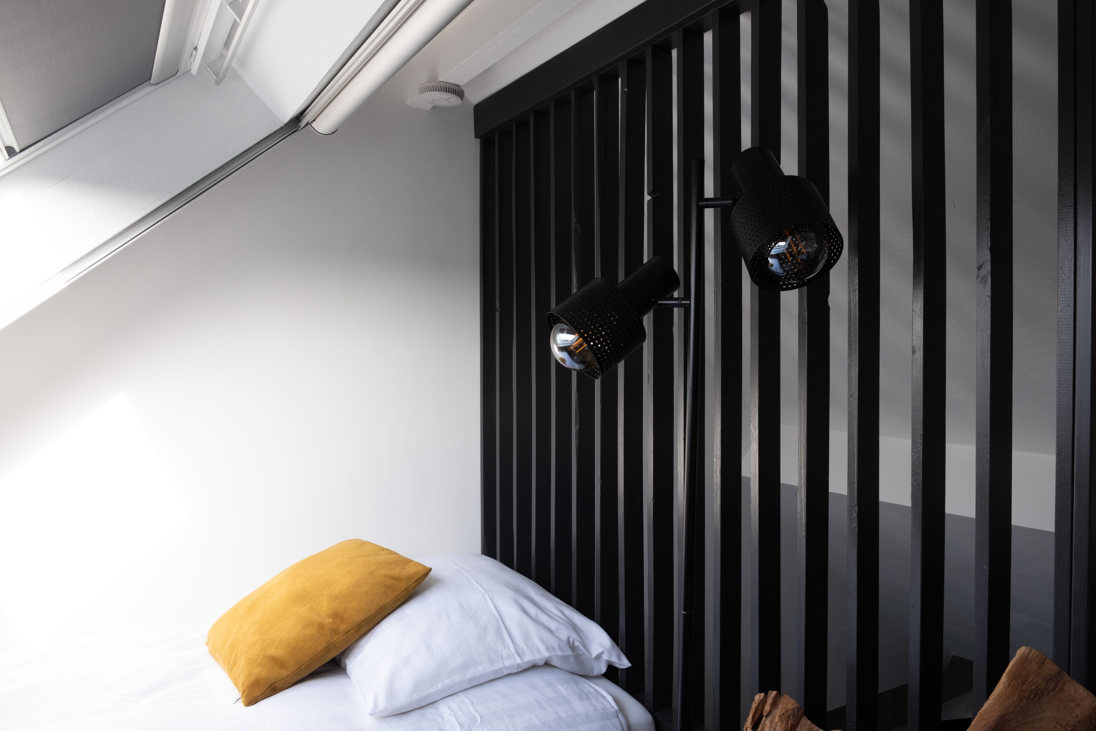 A bedroom with a bed, two pillows, a black wall-mounted lamp, and a window with blinds.