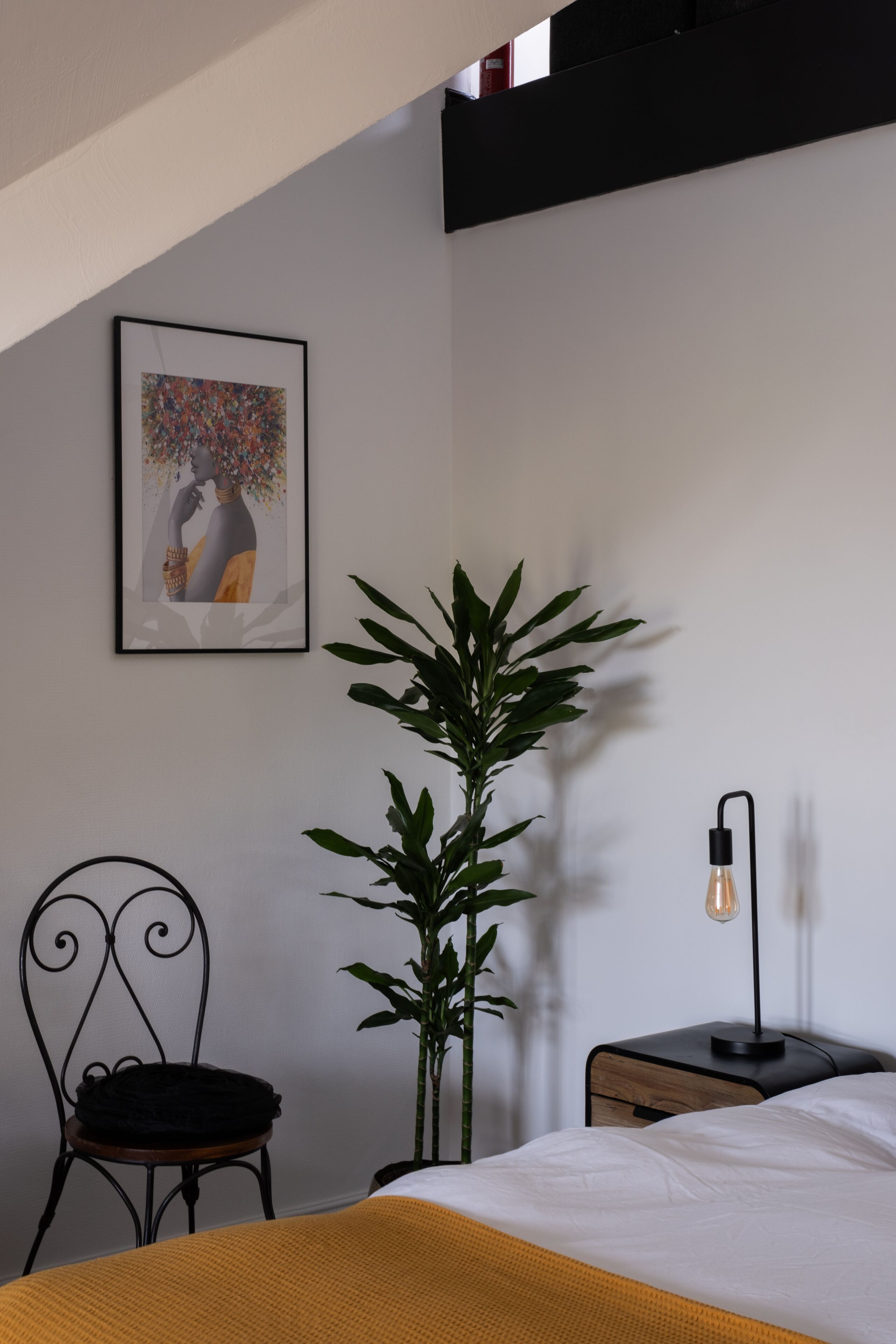 Un dormitorio con cama, una silla, una planta en maceta, una mesita de noche con lámpara y un cuadro en la pared.