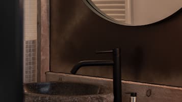 A bathroom with a round mirror, a black faucet, a stone basin, a bottle of shampoo, and a glass of water.