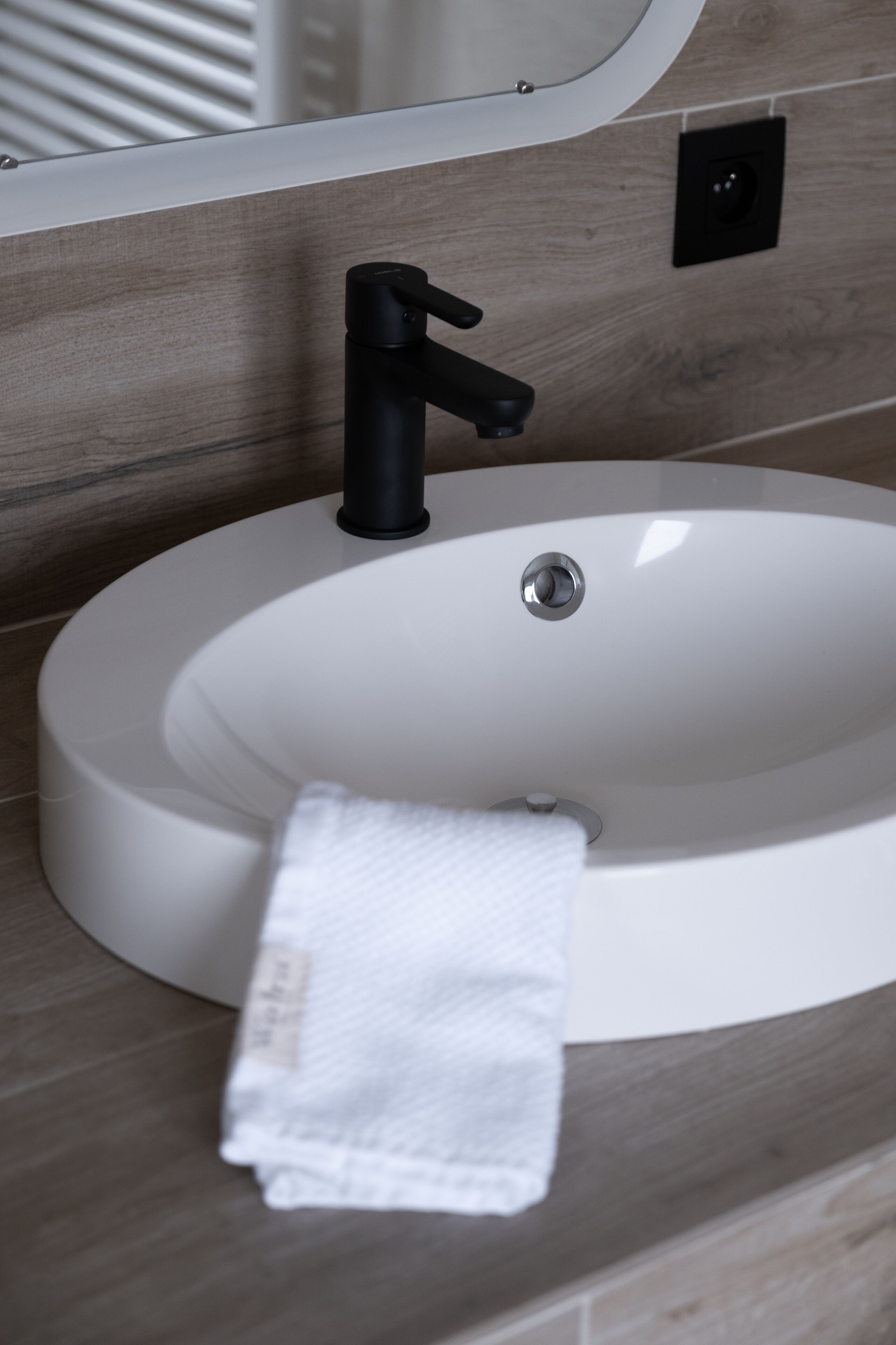 Un lavabo blanc dans une salle de bain, avec un robinet noir, une serviette blanche accrochée sur le côté et un miroir au-dessus du lavabo.