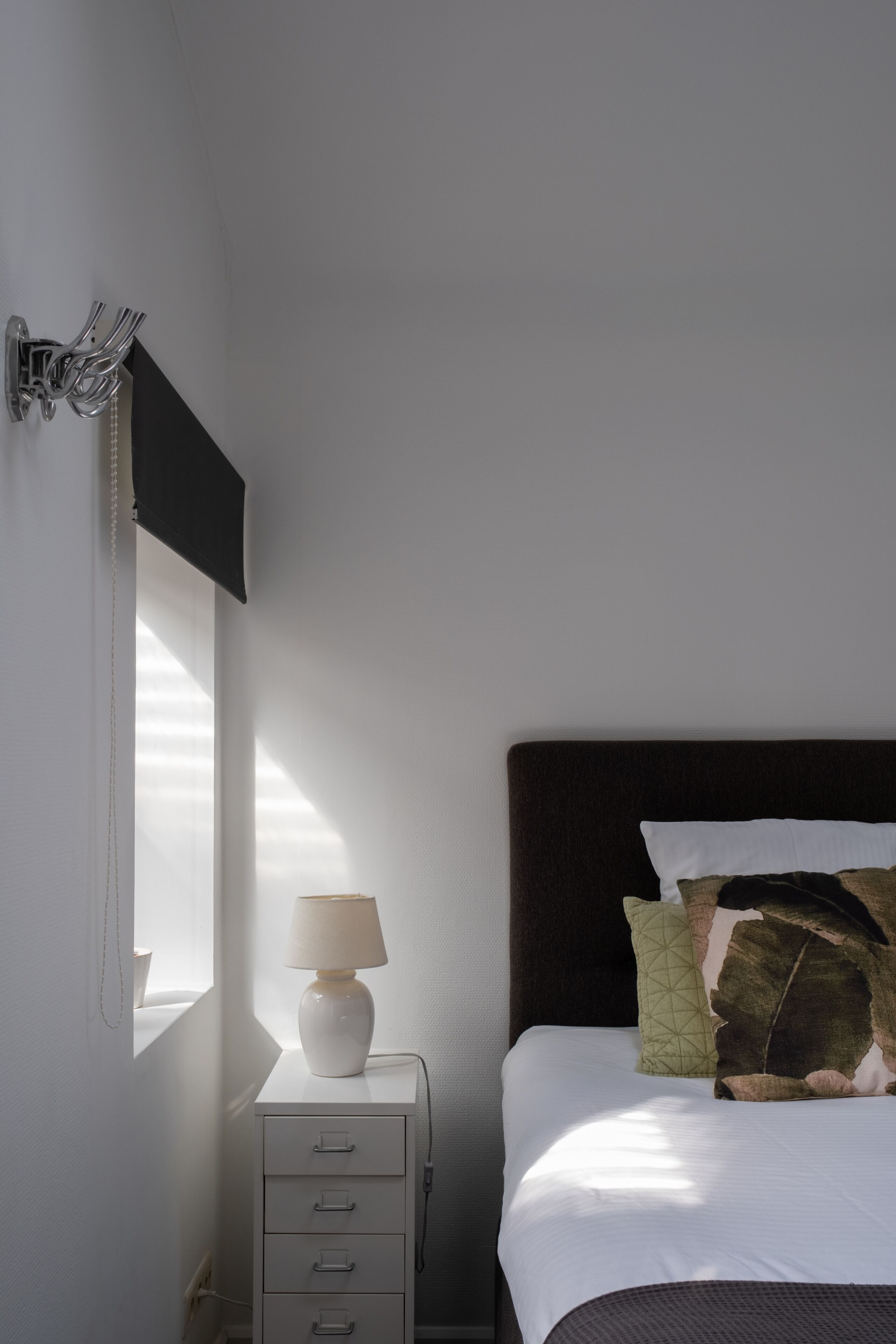 A neatly made bed with a dark headboard, a bedside lamp, and a small white nightstand with drawers.