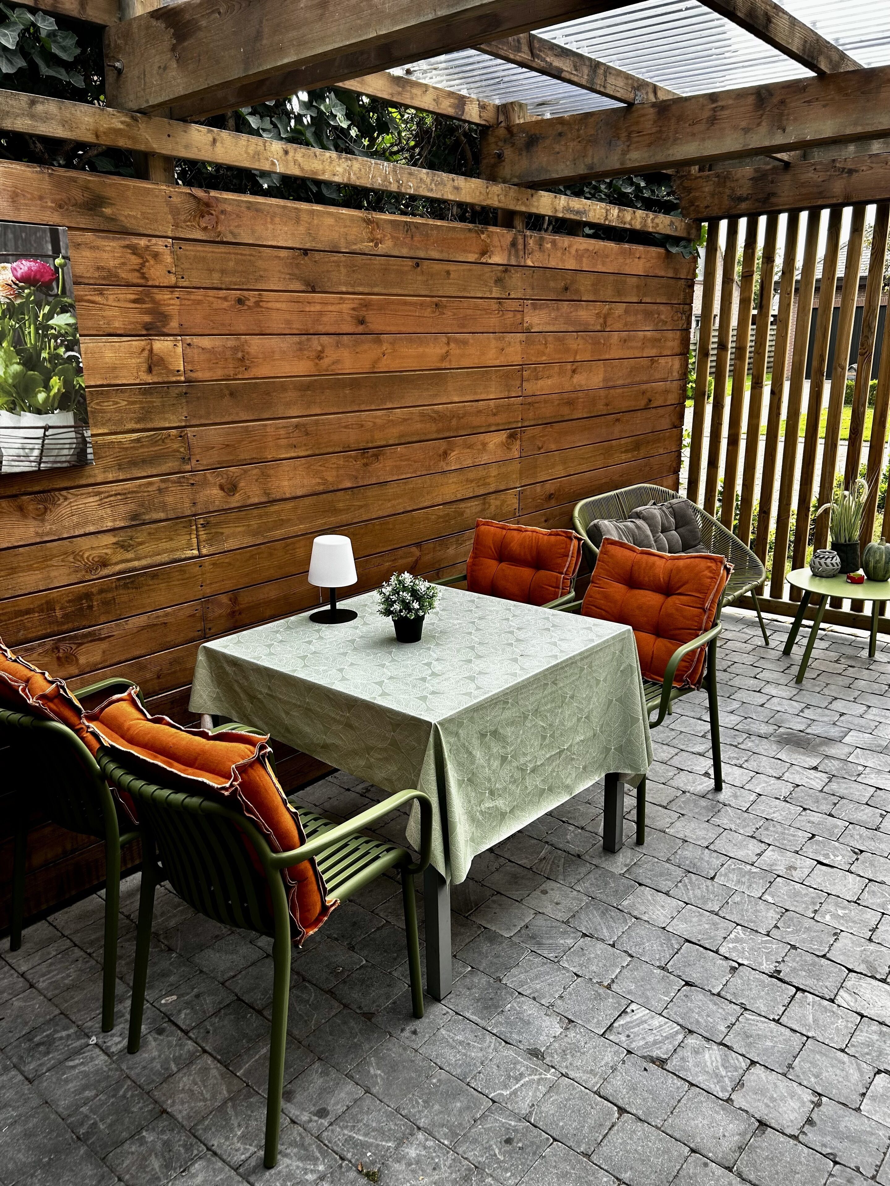 An outdoor patio with a wooden pergola, a table with a white tablecloth, and green and orange cushioned chairs.