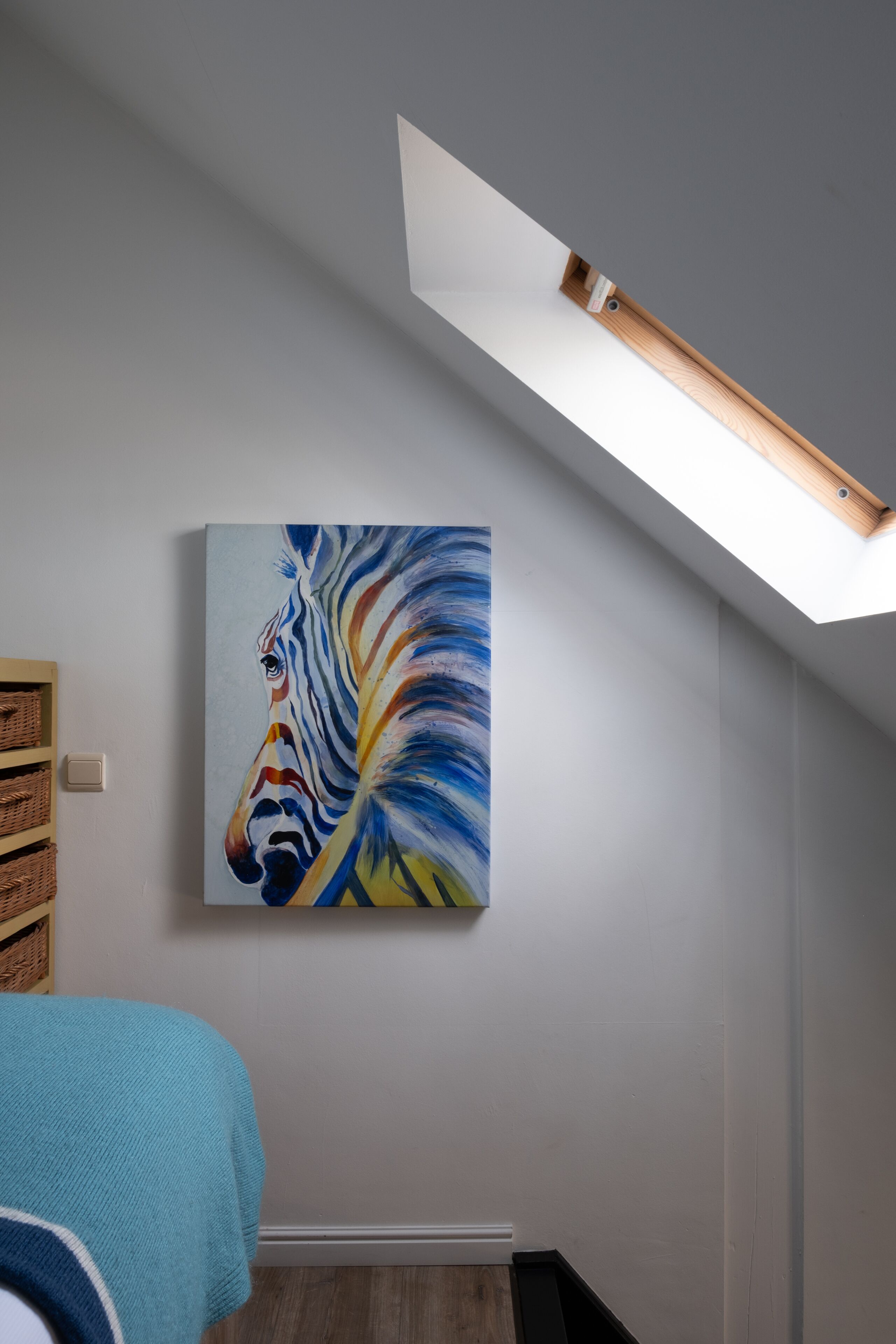 Une pièce avec une lucarne, un tableau représentant une zèbre, une étagère en bois et une couverture bleue.