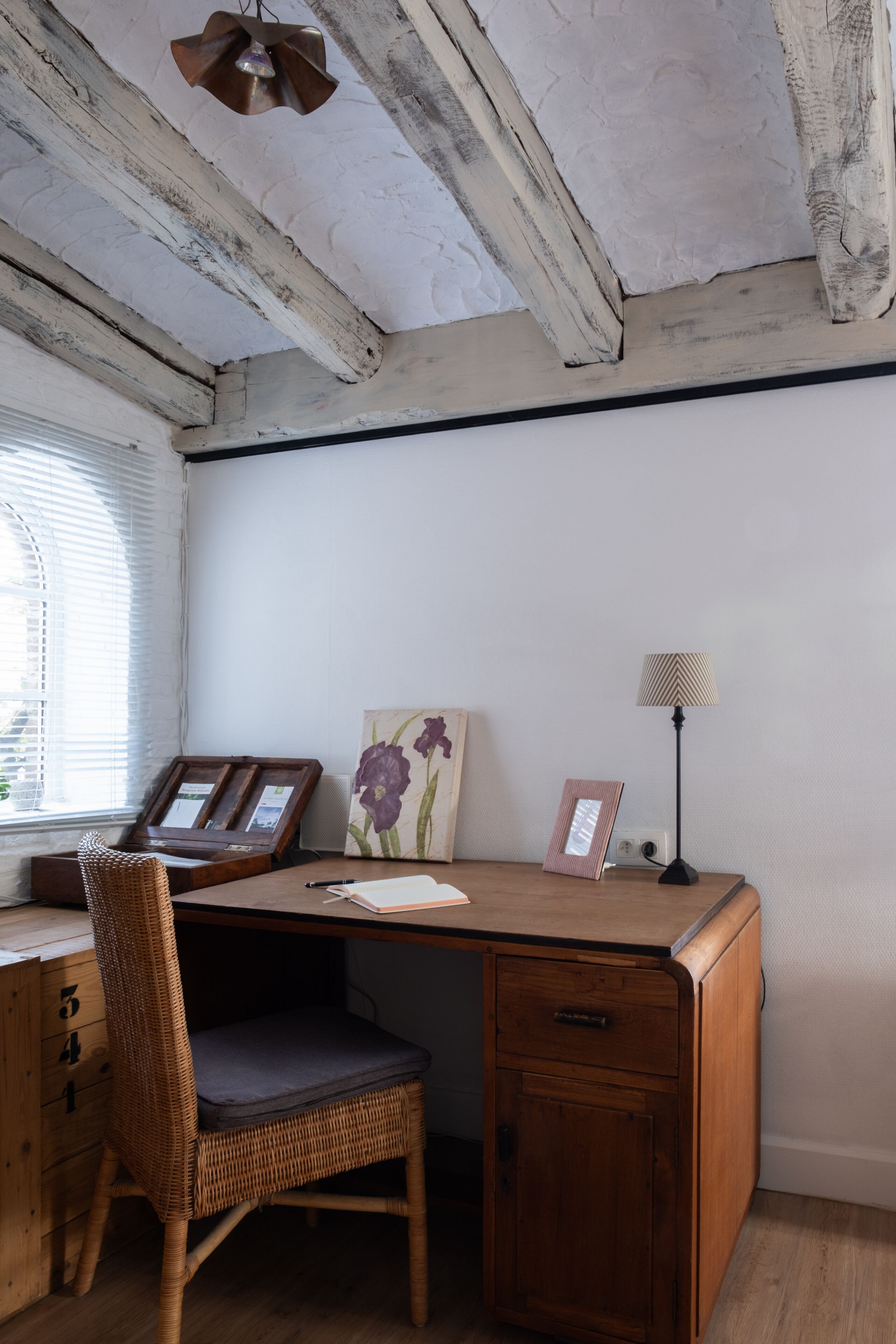 Ein Holztisch mit einem Korbstuhl, einer Lampe und einem gerahmten Bild an der Wand.