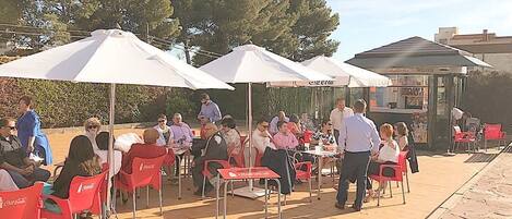 An outdoor dining area with red chairs and white umbrellas, a Coca-Cola branded table, and a building with a sign that reads "Coca-Cola Restaurant" in the background.