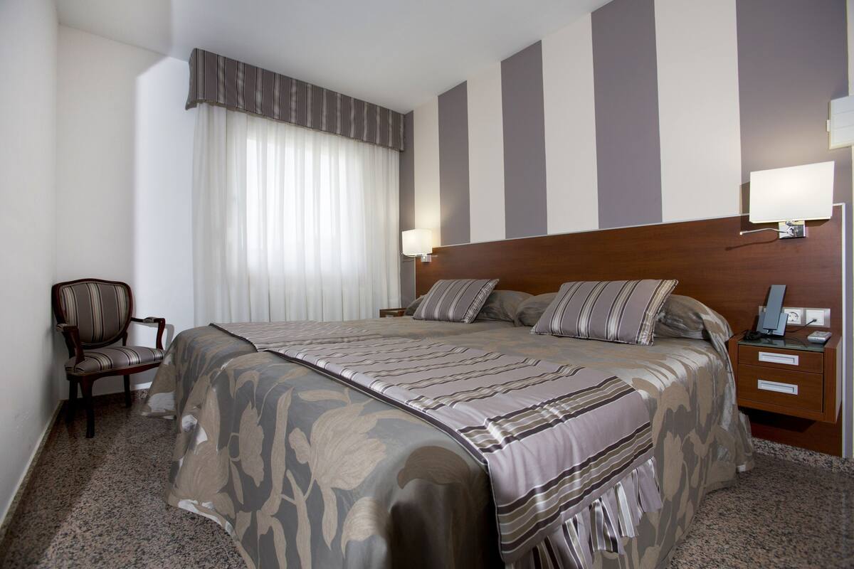 A hotel room with a large bed, a wooden nightstand, a chair, and a window with curtains.