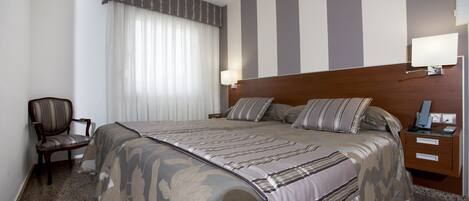 A hotel room with a large bed, a wooden nightstand, a chair, and a window with curtains.