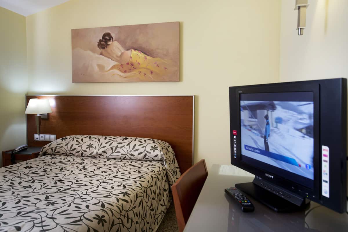 A hotel room with a bed, a television, a painting on the wall, and a nightstand with a lamp.
