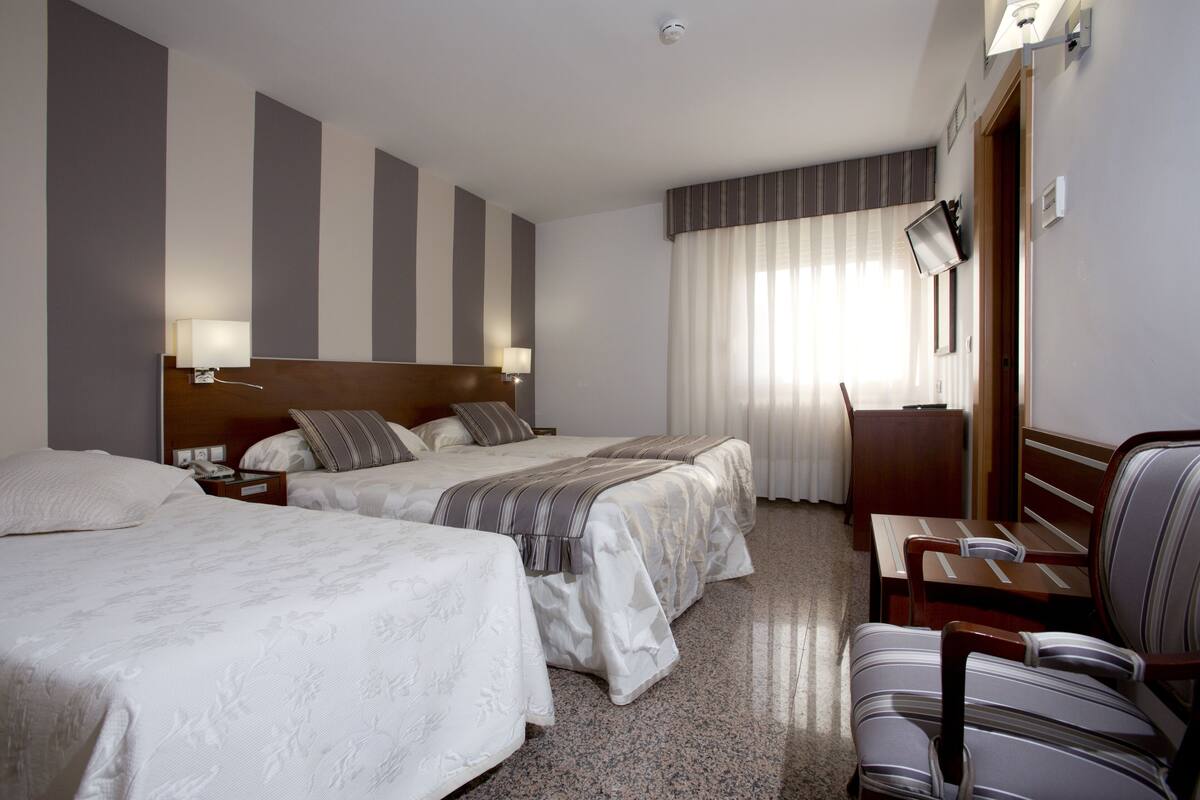 A hotel room with two beds, a desk, a chair, and a large window with curtains.