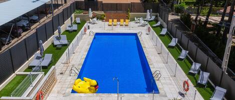 Piscina rectangular con interior azul, rodeada por una cerca. Hay sillas de descanso y sombrillas a lo largo de la piscina.