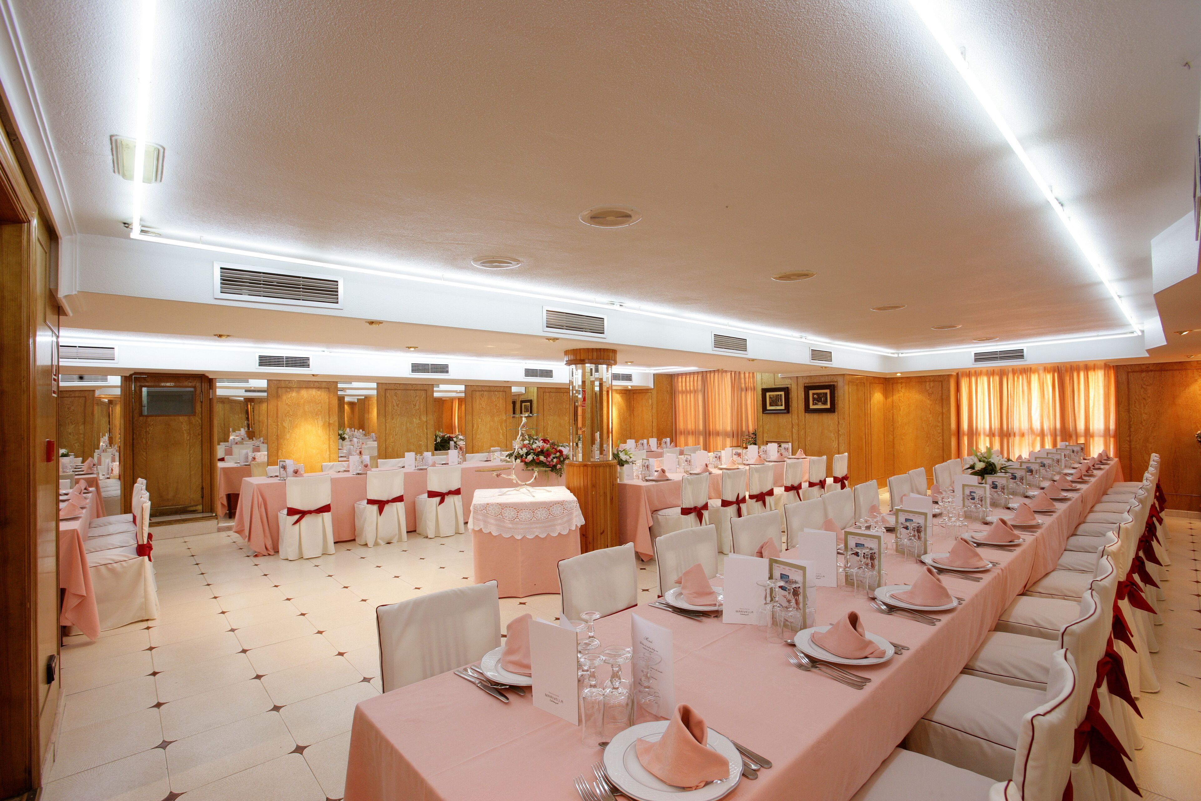 Un salón de banquetes con largas mesas dispuestas para un evento formal, con manteles blancos y rosas, sillas blancas con cintas rojas y una araña de cristal.