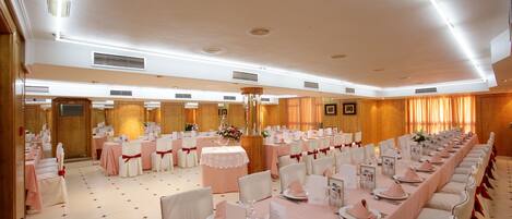 Un salón de banquetes con largas mesas dispuestas para un evento formal, con manteles blancos y rosas, sillas blancas con cintas rojas y una araña de cristal.